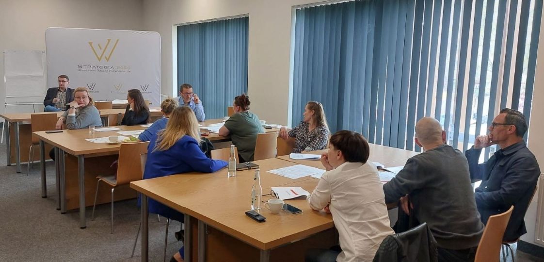 Zdjęcie przedstawia uczestników warsztatu, zgromadzonych w sali konferencyjnej. Osoby siedzą przy stolikach. Na stołach rozłożone są różne dokumenty. W lewym górnym rogu przy stole siedzi Pan prowadzący przodem do stolików z uczestnikami, za nim stoi tablica przedstawiająca logo Strategii WrOF 2050.