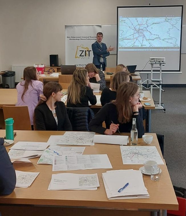 Zdjęcie przedstawia uczestników warsztatu, zgromadzonych w sali konferenycjnej. Osoby siedzą przy stolikach. U góry zdjęcia stoi Pan. który prezentuje mapę wyświetlaną na ekranie. Ekran znajduje się z prawej strony prowadzącego, natomiast z lewej strony za prezenterem stoi rollup z logo ZIT WrOF. Logo znajduje się na białym tle, żółty obrys nawiązujący do obszaru WrOF zawiera w środku czarny napis: ZIT.